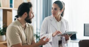 Female doctor in a white coat consulting with a male patient, using a tablet to review medical records.